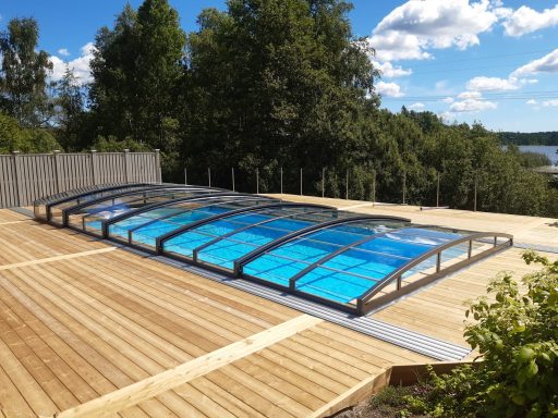 Piscine couverte avec un fond bleu, entourée d'une terrasse en bois et de verdure.
