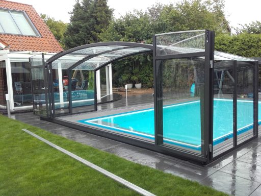 Piscine avec un abri transparent et terrasse en pavés, entourée de verdure.