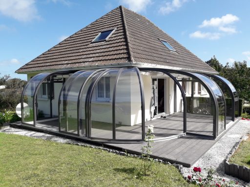 Maison moderne avec véranda en verre et toiture à double pente, jardin soigné.