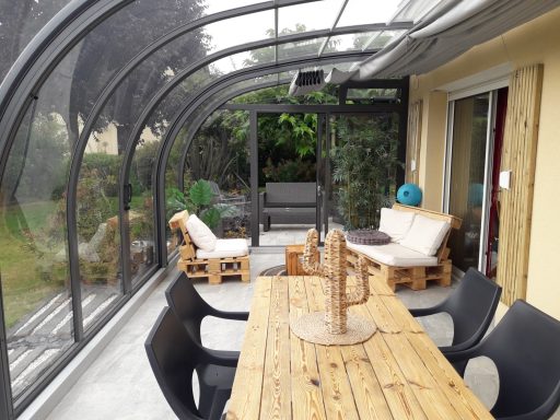 Véranda avec une grande table en bois et des fauteuils confortables, entourée de verdure.