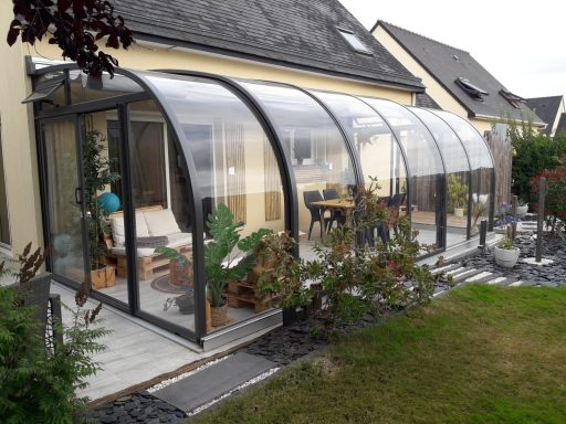 Véranda en verre avec mobilier, entourée de plantes et d’un jardin.