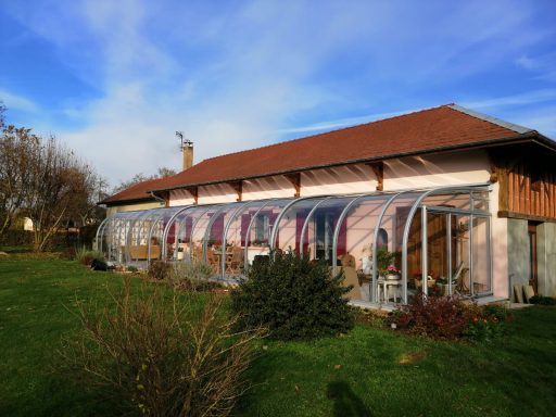 Maison avec véranda en verre, jardin verdoyant et ciel bleu.